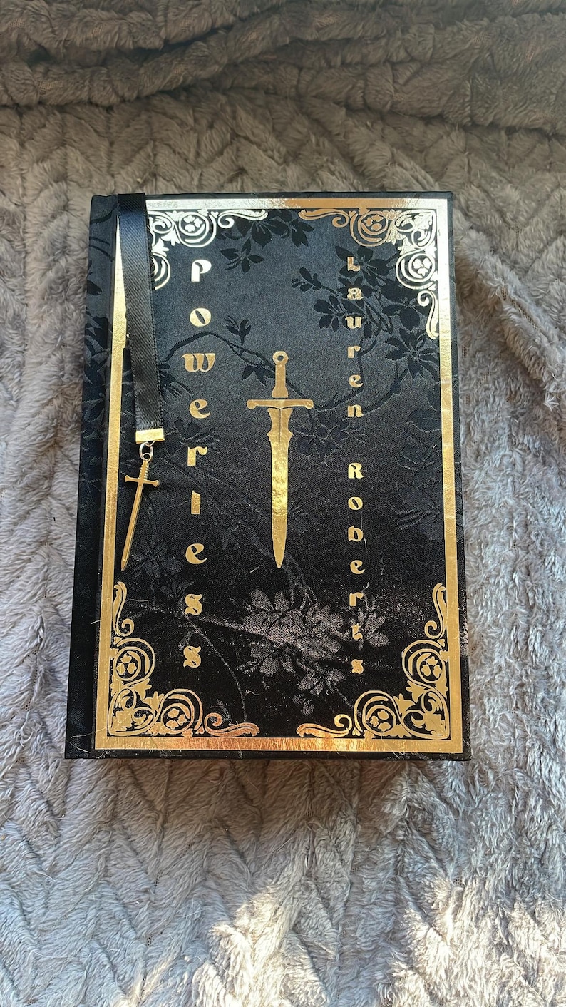 May include: A black book with a gold-coloured design and the title "Powerless" by Lauren Roberts. A gold-coloured sword and cross are on the cover.