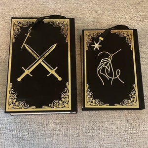 May include: Two black hardcover books with gold accents and decorative designs. The book on the left has a gold sword design and the book on the right has a gold star and needle design.