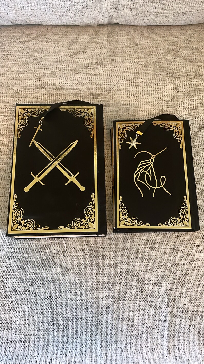 May include: Two black hardcover books with gold accents and decorative designs. The book on the left has a gold sword design and the book on the right has a gold star and needle design.