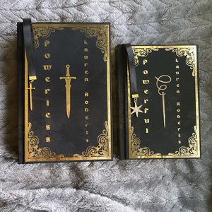 May include: Two black hardcover books with gold accents and decorative designs. The books have black ribbon bookmarks with metal charms. The book on the left has a sword charm and the text "Powerless" on the cover. The book on the right has a star charm and the text "Powerful" on the cover.