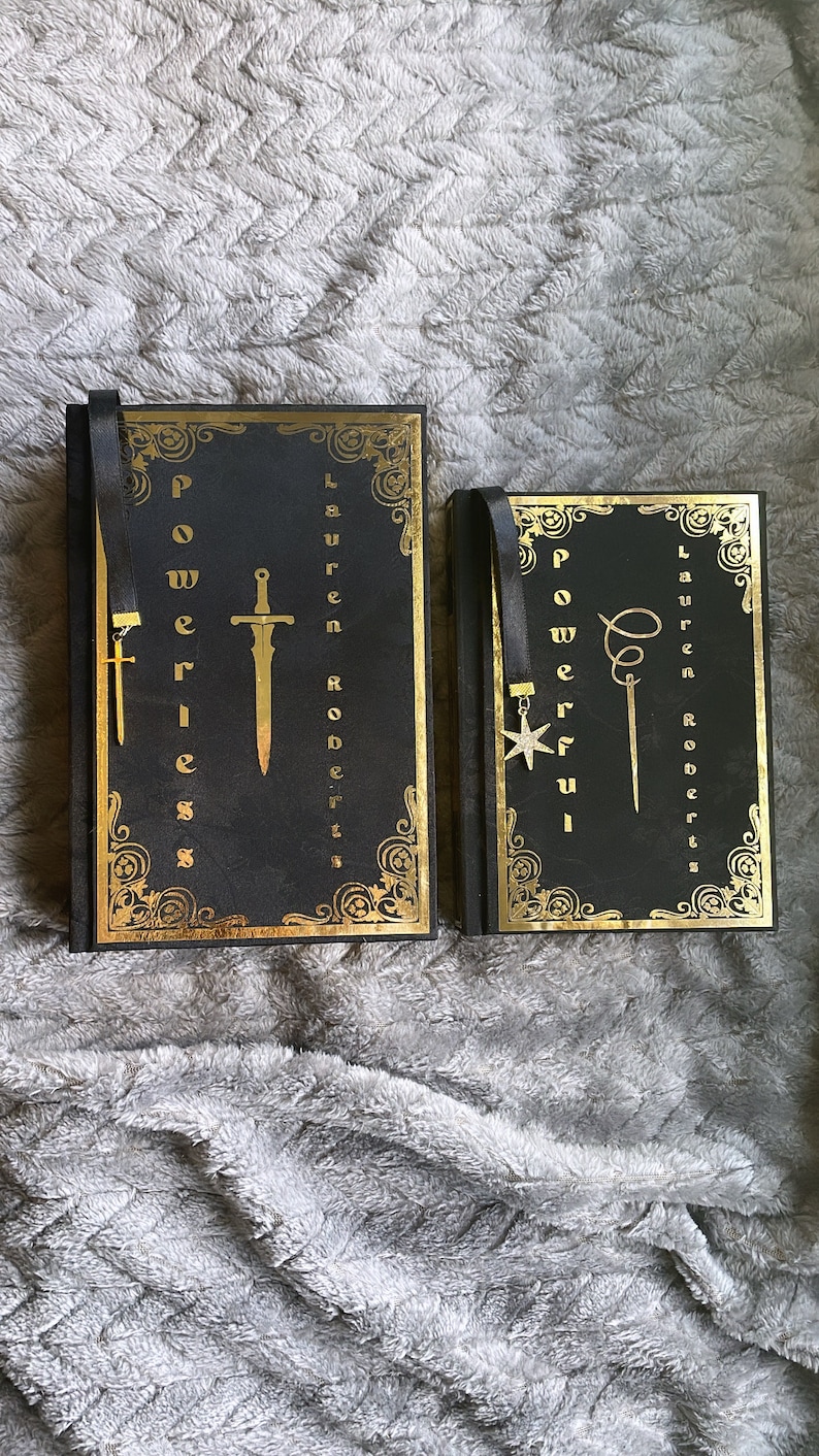 May include: Two black hardcover books with gold accents and decorative designs. The books have black ribbon bookmarks with metal charms. The book on the left has a sword charm and the text "Powerless" on the cover. The book on the right has a star charm and the text "Powerful" on the cover.