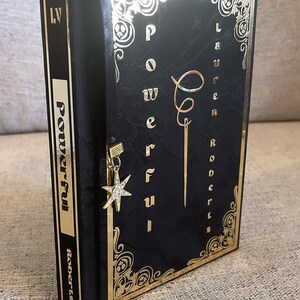 May include: A black book with gold lettering and a gold bookmark with a star charm. The book title is "Powerful" by Lauren Roberts.
