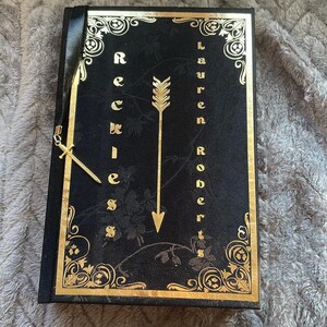 May include: A black journal with a gold embossed design featuring an arrow and the words "Reckless" and "Lauren Roberts".