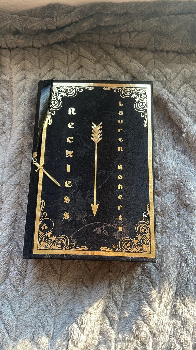 May include: A black journal with a gold embossed design featuring an arrow and the words "Reckless" and "Lauren Roberts".