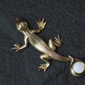 May include: A gold-toned metal lizard door knocker with a white porcelain button. The lizard has a detailed design and is mounted on a black surface.