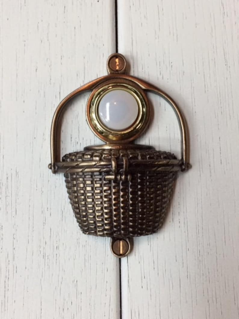 May include: A bronze-toned cabinet pull in the shape of a basket. The pull has a woven basket design with a handle and a circular, light-colored stone set in a gold-toned frame. The pull is mounted on a white cabinet door.