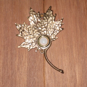 May include: A decorative brass door knocker in the shape of a maple leaf. The leaf has a textured surface and a central, round, opalescent stone. The knocker is mounted on a wooden door.