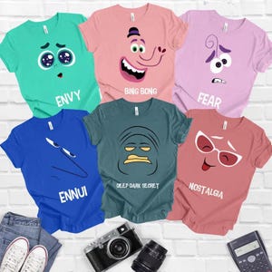 Op de afbeelding: Zes verschillende T-shirts met personages uit de film Inside Out. Elk shirt heeft een ander personage en emotie, waaronder Joy, Sadness, Fear, Anger, Disgust en Bing Bong. De shirts zijn allemaal in verschillende kleuren, waaronder roze, blauw, groen en paars.
