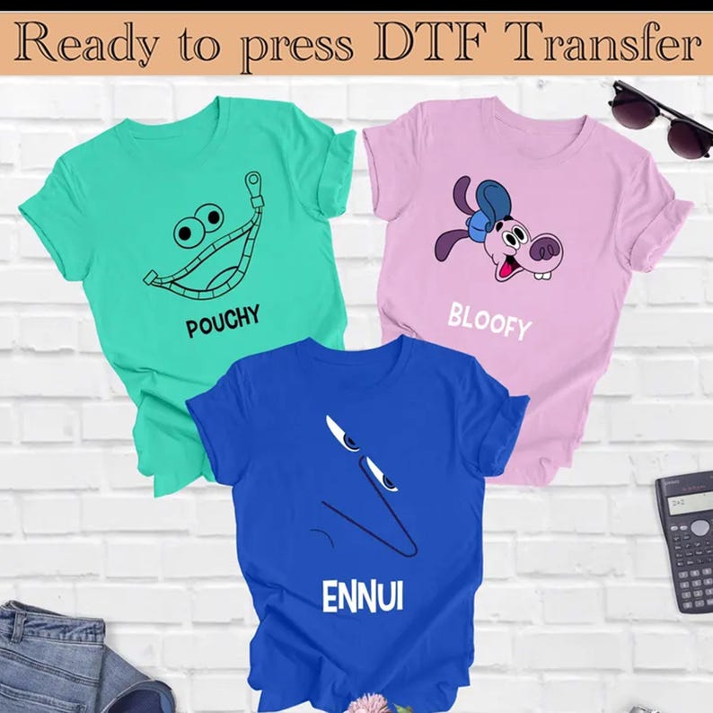 Op de afbeelding: Drie T-shirts met verschillende ontwerpen. Het eerste T-shirt is groen met een cartoon gezicht en de tekst "POUCHY". Het tweede T-shirt is roze met een cartoon hond en de tekst "BLOOFY". Het derde T-shirt is blauw met een cartoon gezicht en de tekst "ENNUI".