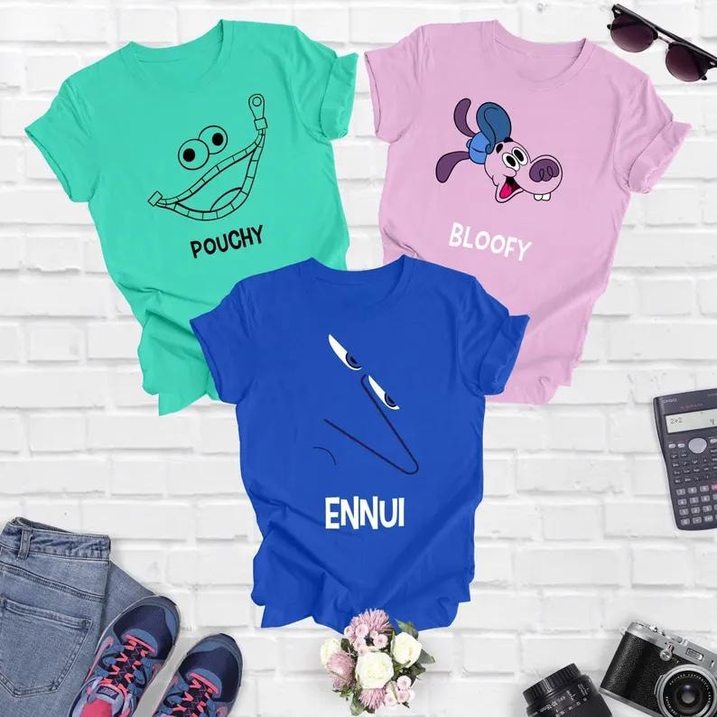 Op de afbeelding: Drie T-shirts met verschillende cartoonfiguren en tekst. Het eerste T-shirt is turquoise met een groene ritsmond en de tekst "POUCHY". Het tweede T-shirt is roze met een cartoonhond en de tekst "BLOOFY". Het derde T-shirt is blauw met een cartoongezicht en de tekst "ENNUI".