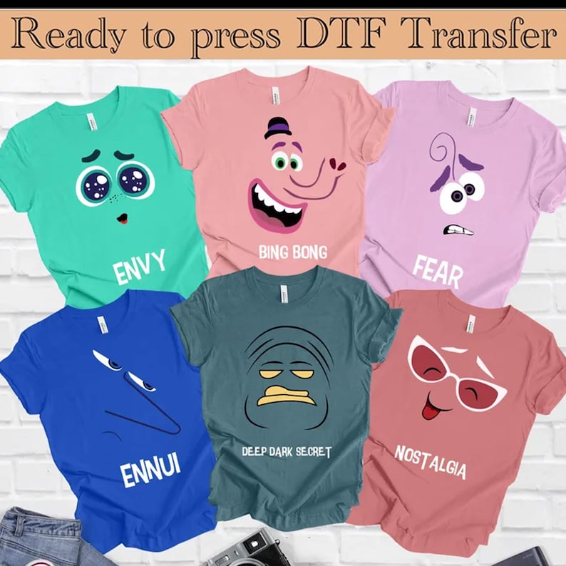 Op de afbeelding: Vijf verschillende gekleurde T-shirts met cartoon gezichten en tekst. De shirts zijn teal, roze, lichtblauw, donkerblauw en koraal. De tekst op de shirts luidt "Envy", "Bing Bong", "Fear", "Ennui", "Deep Dark Secret" en "Nostalgia".