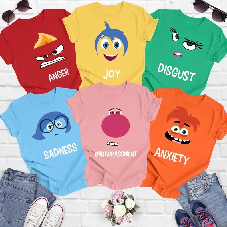 Op de afbeelding: Zes kleurrijke T-shirts met personages uit de film Inside Out. De shirts zijn rood, geel, groen, blauw, roze en oranje. Elk shirt heeft het gezicht van een ander personage erop, met de naam van het personage eronder. De personages zijn Woede, Vreugde, Afkeer, Verdriet, Schaamte en Angst.