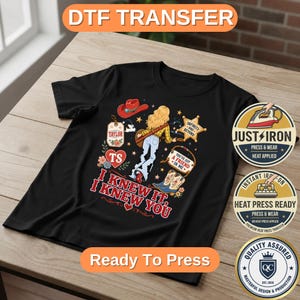May include: A black t-shirt featuring a graphic print of a person with blonde hair, a red cowboy hat, blue jeans, and cowboy boots. The design includes a star, a heart, and text that reads "I KNEW IT, I KNEW YOU", "TAYLOR", "TS", and "YOU'VE GOT A FRIEND IN ME".