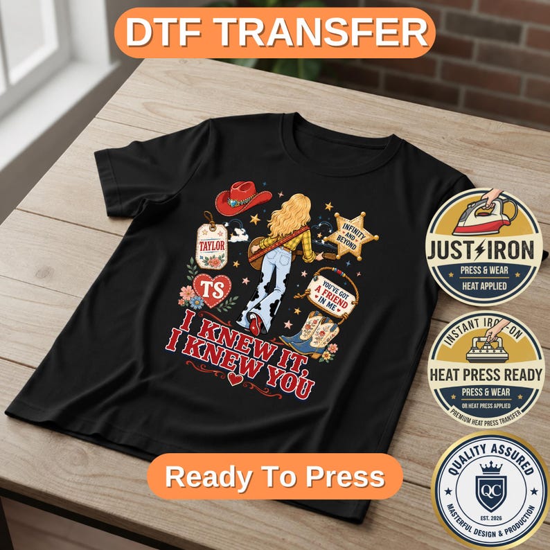 May include: A black t-shirt featuring a graphic print of a person with blonde hair, a red cowboy hat, blue jeans, and cowboy boots. The design includes a star, a heart, and text that reads "I KNEW IT, I KNEW YOU", "TAYLOR", "TS", and "YOU'VE GOT A FRIEND IN ME".