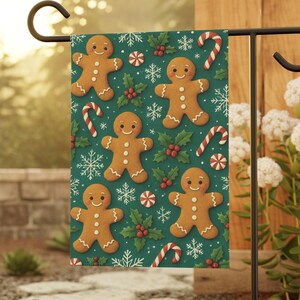May include: A festive garden flag featuring a repeating pattern of gingerbread men, candy canes, snowflakes, and holly berries on a teal background. The flag is displayed on a black metal stand in an outdoor setting.