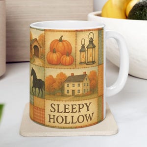 May include: White ceramic mug featuring a patchwork design with autumn-themed illustrations. The design includes pumpkins, a covered bridge, lanterns, a house, and a horse. The text "SLEEPY HOLLOW" is displayed on the mug.