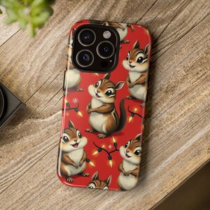 May include: A red phone case featuring a repeating pattern of cartoon chipmunks. The chipmunks are brown and white with smiling faces. The case also has a string of yellow lights and snowflakes. The phone case is on a wooden surface.