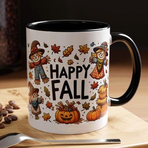 May include: White ceramic mug with a black interior and handle, featuring a "Happy Fall" design. The design includes scarecrows, pumpkins, and autumn leaves. A spoon and a pastry are in the foreground.