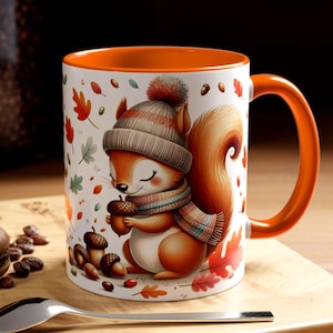 May include: White ceramic mug with an orange interior and handle. The mug features a cartoon squirrel wearing a hat and scarf, holding an acorn. Autumn leaves and acorns decorate the mug's exterior. A spoon and a macaron are in the foreground.
