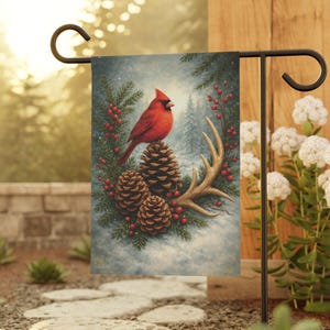 Puede incluir: Una bandera de jardín con un cardenal rojo brillante posado sobre piñas y cornamentas, rodeado de ramas de hoja perenne y bayas rojas. La escena invernal se desarrolla sobre un fondo de bosque nevado, creando una decoración festiva para exteriores.