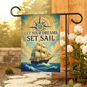 May include: A garden flag featuring a vintage illustration of a sailing ship on the ocean, with a compass rose and the text "LET YOUR DREAMS SET SAIL" against a cloudy sky background. The flag is displayed on a black metal stand.