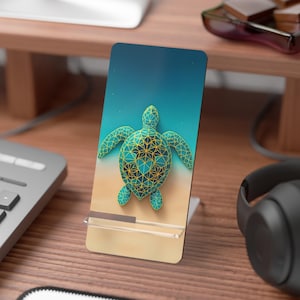 May include: A teal and gold geometric sea turtle design on a phone stand. The stand is clear with a rectangular base and a slot to hold a phone. The background is a gradient of blue and tan.