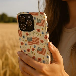 May include: A close-up of a white iPhone case with a pattern of red, white, and blue American flags and flowers. The case is being held by a person's hand.