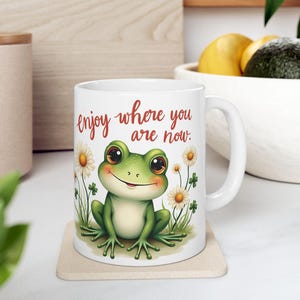 May include: White ceramic mug with a green frog illustration and the text "enjoy where you are now." The frog is surrounded by white daisies and green leaves.