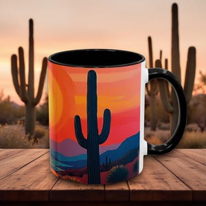 May include: A white ceramic mug with a black rim and handle. The mug features a colorful illustration of a desert landscape with a large cactus in the foreground and a sunset in the background.