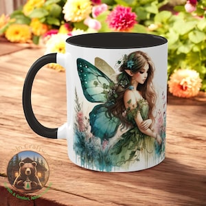 May include: White ceramic mug with a black handle and interior. The mug features a watercolor illustration of a fairy with teal wings and a green dress. The mug is decorated with flowers and foliage.