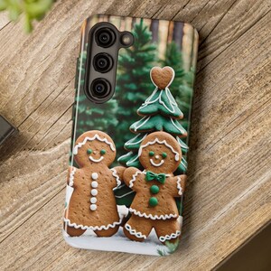 May include: A phone case with a gingerbread man and woman design. The gingerbread people are standing in front of a green Christmas tree with a heart-shaped ornament. The background is a blurry image of a forest.