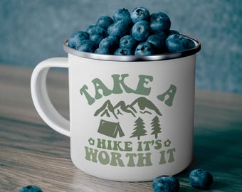 May include: White enamel camping mug filled with blueberries. The mug has a silver rim and a handle. The text "TAKE A HIKE IT'S WORTH IT" is printed in green. Blueberries are scattered around the mug on a wooden surface.