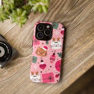 May include: Pink phone case with a Christmas-themed pattern. The design features cartoon cats wearing reindeer antlers and Santa hats, along with wrapped gifts, ornaments, and snowflakes. The case is on a wooden surface.