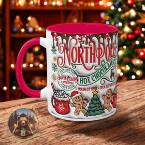May include: White ceramic mug with a red rim and handle. The mug features a festive design with the words "North Pole Hot Chocolate Co." and illustrations of gingerbread people, a Christmas tree, and a cup of hot chocolate with marshmallows. The text "Authentic Santa Claus Approved Since 1891" and "Warm Up Here Open All Winter" are also included.