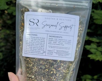 May include: A clear plastic bag filled with a herbal tea blend. The label reads "Seasonal Support" and lists ingredients like Nettle Leaf and Peppermint Leaf. The bag is held by a hand against a blurred green background.