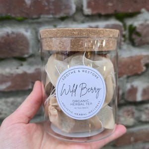 May include: A clear glass jar with a cork lid, filled with tea bags. A white label on the jar reads "Wild Berry Organic Herbal Tea" with the text "Soothe & Restore" and "Holistic Herbal Wellness" around the edges. The jar is held in front of a brick wall.