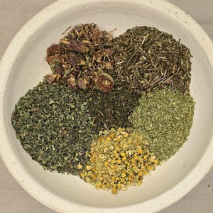 May include: A white ceramic bowl filled with various dried herbs. The herbs are arranged in sections, showcasing different colors and textures, including green, brown, and yellow. The bowl sits on a neutral-toned surface.