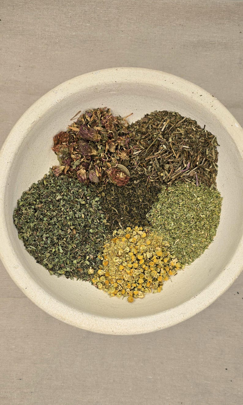 May include: A white ceramic bowl filled with various dried herbs. The herbs are arranged in sections, showcasing different colors and textures, including green, brown, and yellow. The bowl sits on a neutral-toned surface.
