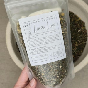 May include: A clear bag of herbal tea blend, "Liver Love", held by a hand. The bag contains dried herbs and flowers. The label includes directions and organic ingredients. The net weight is 48g. The bag is in a bowl.