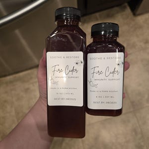 May include: Two glass bottles of Fire Cider Immunity Support, made in a home kitchen. The larger bottle is 16 ounces and the smaller bottle is 8 ounces. Both bottles have a best by date of 08/2025.