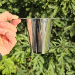 May include: A stainless steel tea infuser held against a backdrop of green foliage. The infuser is cylindrical with a perforated design, featuring a flat rim for resting on a cup. Ideal for steeping loose leaf tea.