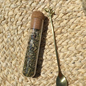 Puede incluir: Un vial de vidrio transparente lleno de hierbas secas, sellado con un tapón de corcho, y una cuchara dorada con un diseño de hoja. La cuchara está junto al vial sobre una superficie beige tejida.