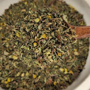 May include: A close-up of a wooden spoon filled with a blend of dried herbs and flowers. The mixture includes green leaves, yellow flower buds, and hints of purple. The spoon is held over a white bowl filled with the same herbal blend.