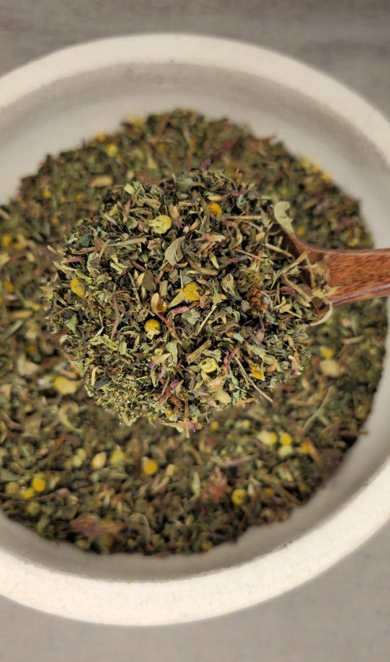 May include: A close-up of a wooden spoon filled with a blend of dried herbs and flowers. The mixture includes green leaves, yellow flower buds, and hints of purple. The spoon is held over a white bowl filled with the same herbal blend.