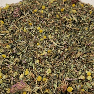 May include: Close-up of a mix of dried herbs and flowers, including yellow chamomile blossoms, green leaves, and purple petals. The blend appears to be a loose-leaf herbal tea, with a variety of textures and colors.