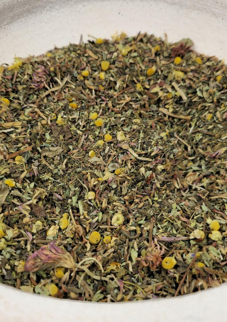 May include: Close-up of a mix of dried herbs and flowers, including yellow chamomile blossoms, green leaves, and purple petals. The blend appears to be a loose-leaf herbal tea, with a variety of textures and colors.