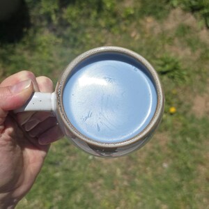 May include: A ceramic mug with a light blue liquid inside. The mug has a white handle and a brown rim. The liquid has a swirl pattern. The mug is held by a person's hand.