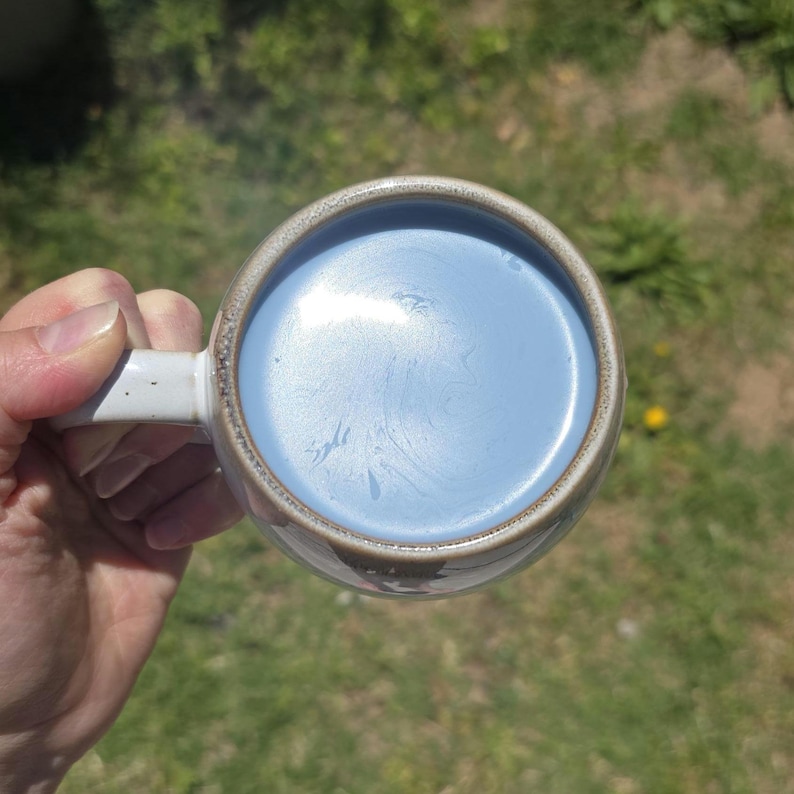 May include: A ceramic mug with a light blue liquid inside. The mug has a white handle and a brown rim. The liquid has a swirl pattern. The mug is held by a person's hand.