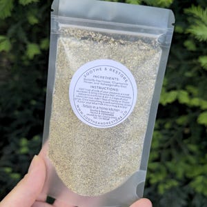 May include: A clear, resealable pouch filled with a light green herbal blend. The pouch is held in a hand and features a circular label with the words "Soothe & Restore" and instructions. The ingredients include Butterfly Pea Flower, Chamomile Flower, and Ashwagandha Root.