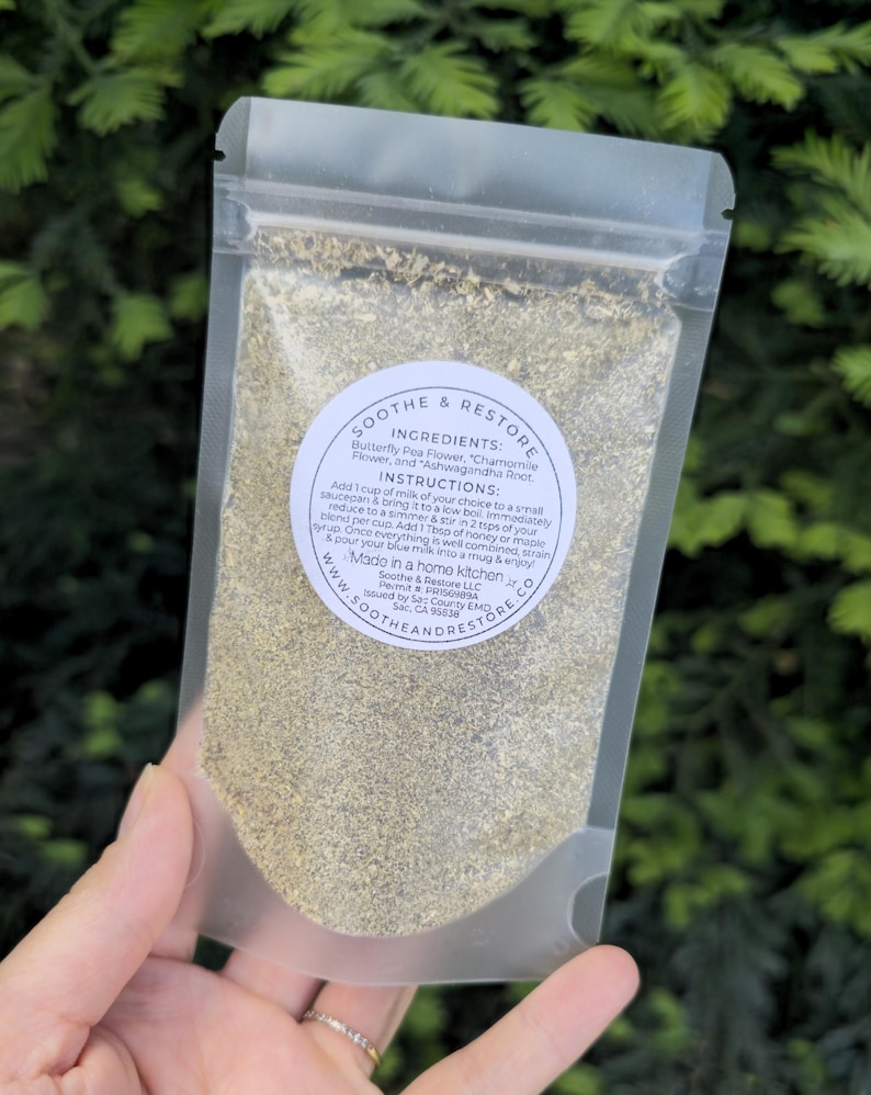 May include: A clear, resealable pouch filled with a light green herbal blend. The pouch is held in a hand and features a circular label with the words "Soothe & Restore" and instructions. The ingredients include Butterfly Pea Flower, Chamomile Flower, and Ashwagandha Root.